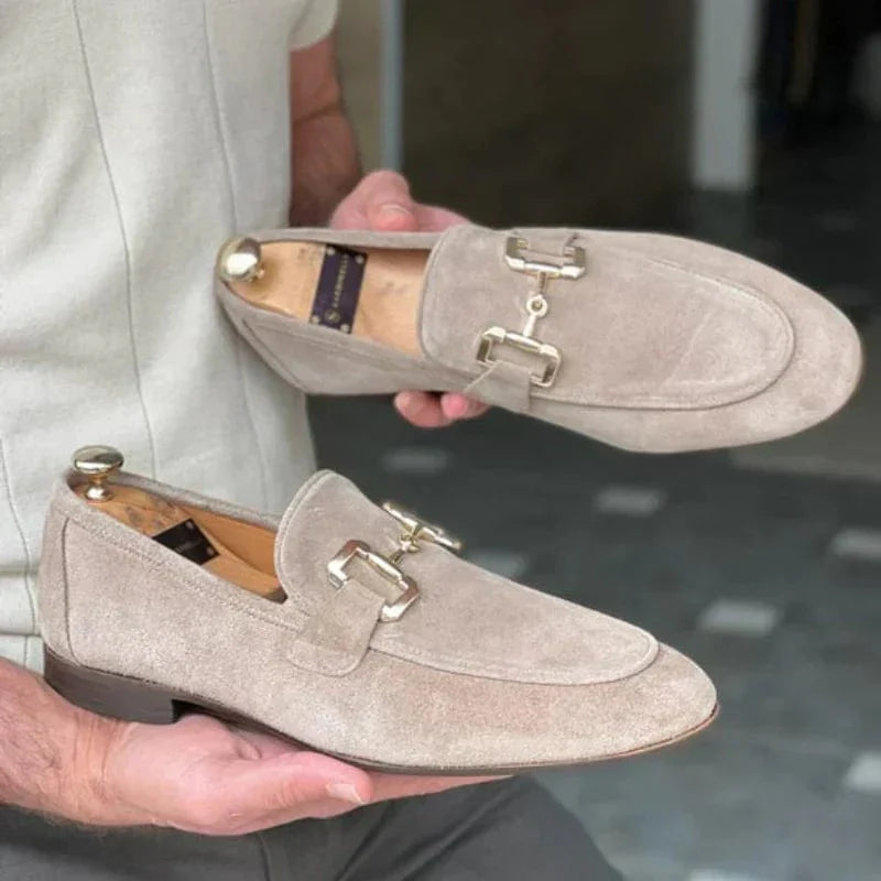 Chaussures mocassins en daim beige pour homme avec boucle dorée, tenues par une personne. Élégance, mode masculine, chaussures de luxe.