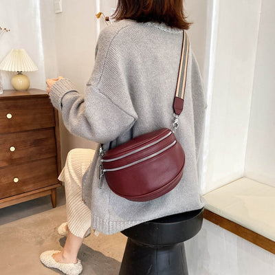 Femme assise avec un pull gris, portant un sac bandoulière bordeaux en cuir, intérieur moderne. Mode automne, accessoires tendance.