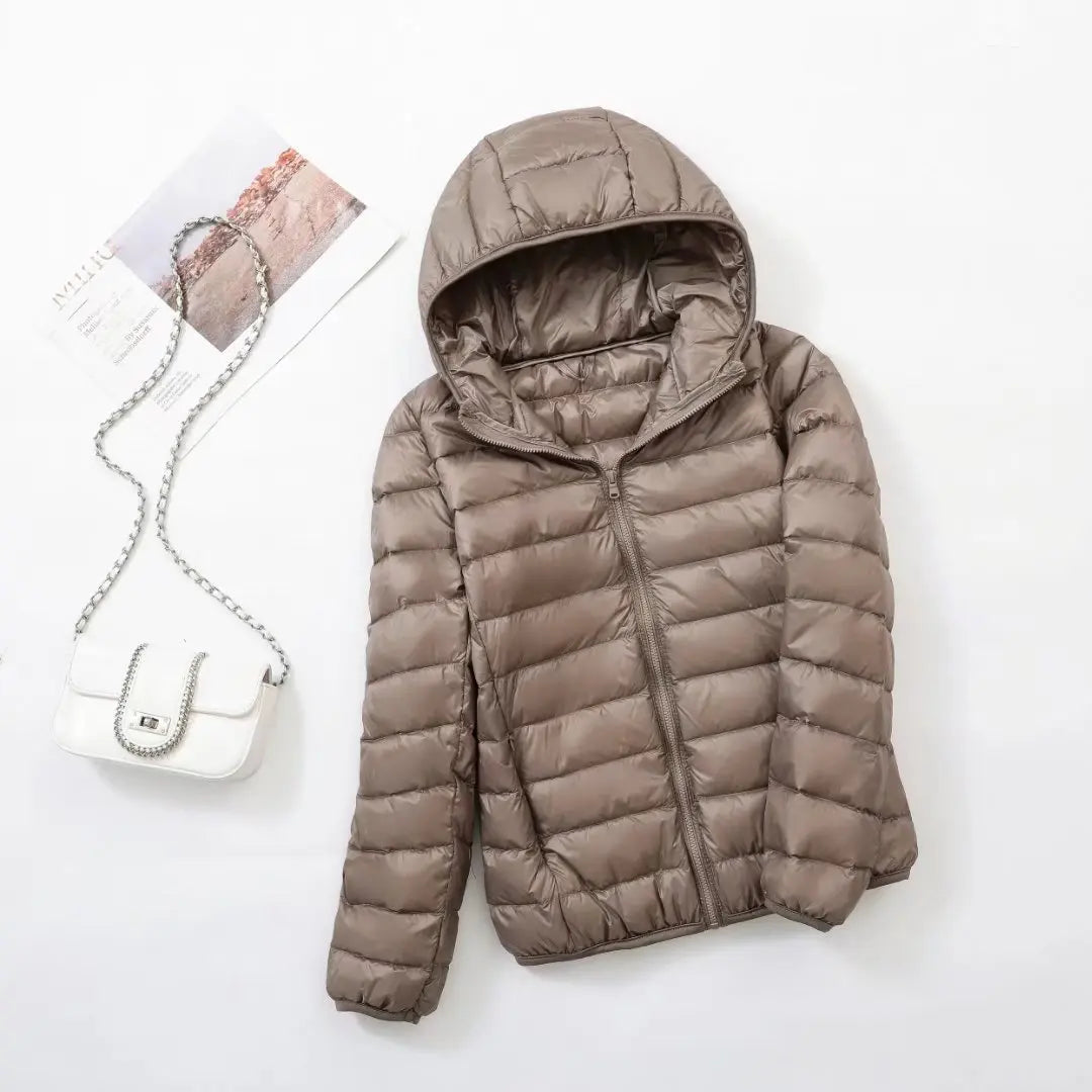 Doudoune marron à capuche posée à plat, accompagnée d'un sac à main blanc avec chaîne argentée, sur fond blanc. Mode hiver, vêtements chauds.