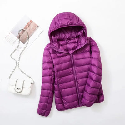 Veste doudoune violette avec capuche sur fond blanc, accompagnée d'un sac à main blanc à chaîne argentée et d'un magazine. Mode hiver femme.