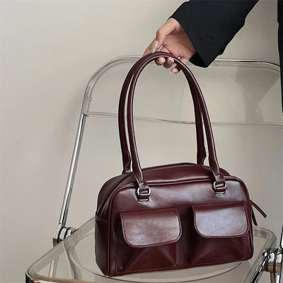 Sac à main en cuir marron foncé avec deux poches avant, posé sur une chaise transparente. Accessoire mode élégant et tendance pour femme.