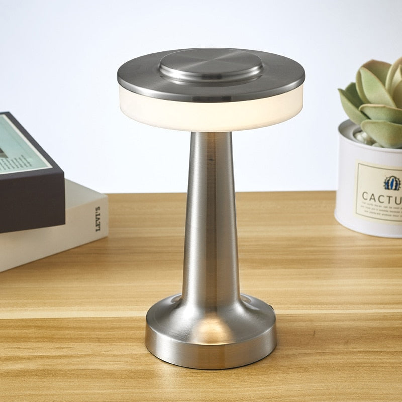 Lampe de table moderne en métal argenté sur bureau en bois, avec livres et plante succulente en arrière-plan. Éclairage LED élégant et contemporain.