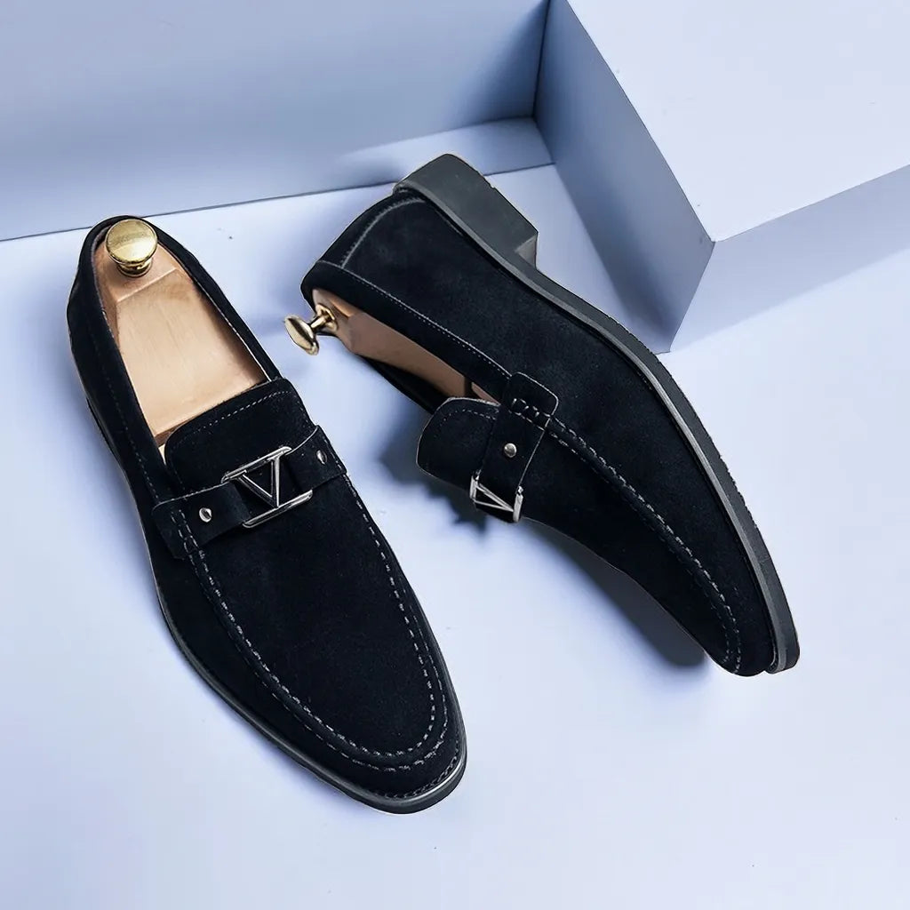 Chaussures mocassins en daim noir pour homme avec boucle métallique, semelle en caoutchouc, posées sur fond gris. Élégance et style moderne.