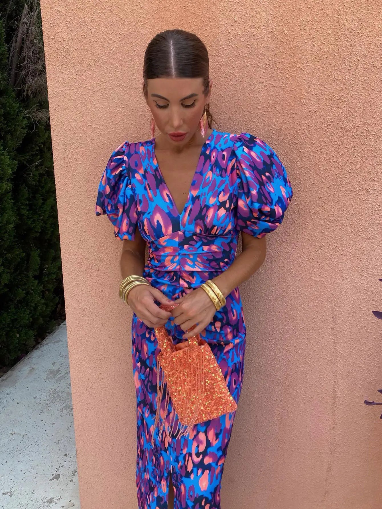 Femme en robe à motifs bleus et roses, manches bouffantes, tenant un sac orange. Fond mur rose. Mode élégante, tenue estivale chic.