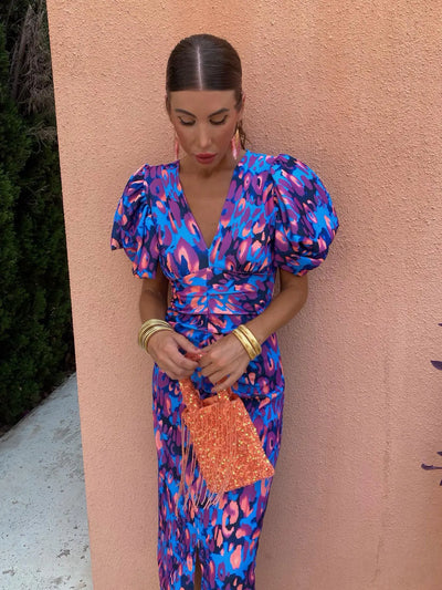 Femme en robe à motifs bleus et roses, manches bouffantes, tenant un sac orange. Fond mur rose. Mode élégante, tenue estivale chic.