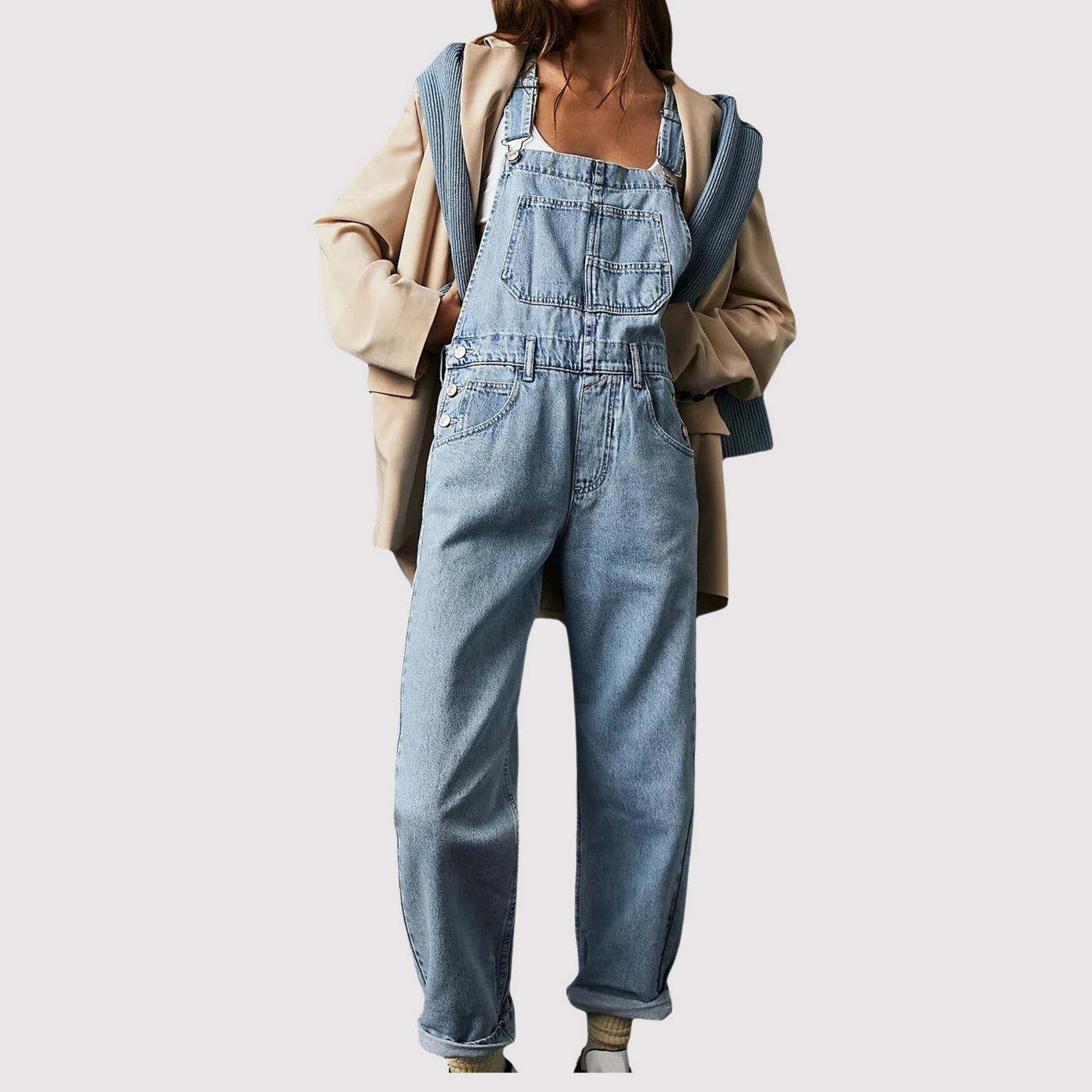 Femme portant une salopette en jean bleu clair, associée à un manteau beige et un pull bleu, mode décontractée, tendance automne-hiver.