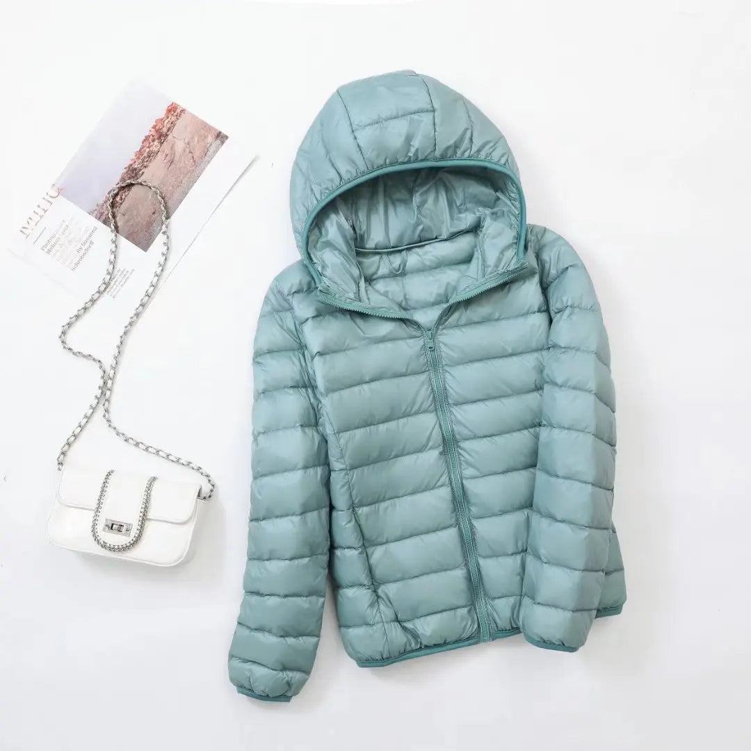 Doudoune légère bleue avec capuche, posée à plat, accompagnée d'un sac à main blanc à chaîne argentée et d'un magazine sur fond blanc.