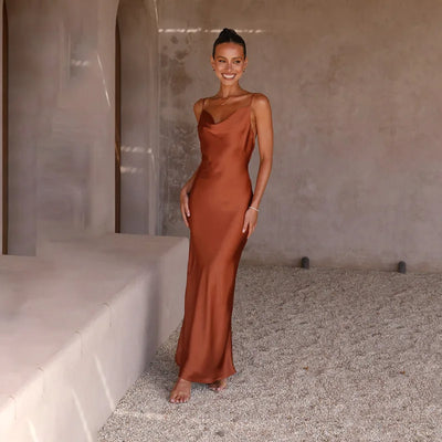 Robe longue en satin marron portée par une femme souriante, debout sur un sol en gravier, devant un mur beige. Mode élégante, tenue de soirée.