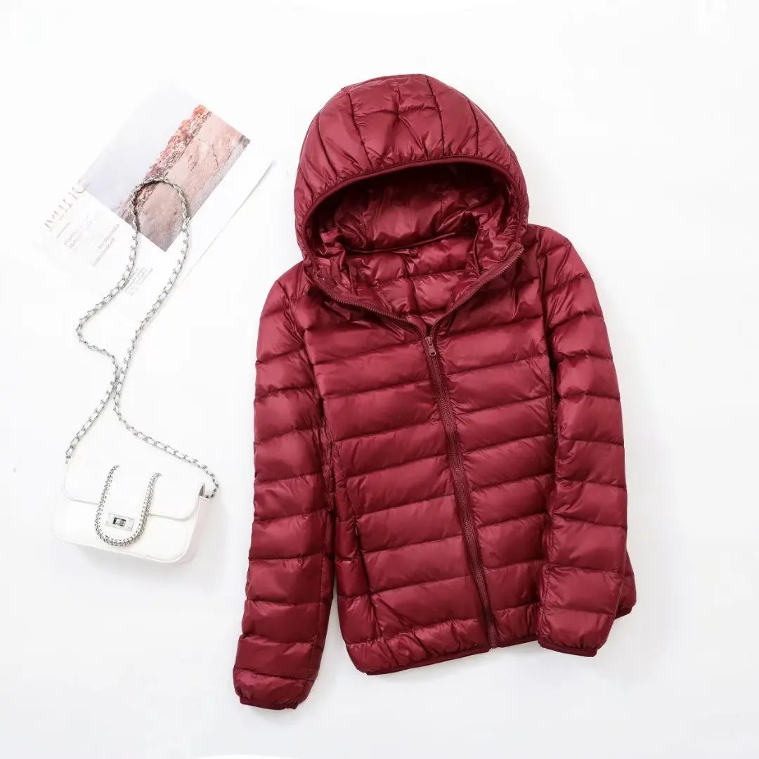 Veste matelassée rouge avec capuche, posée à plat, accompagnée d'un sac à main blanc à chaîne argentée et d'un magazine sur fond blanc.