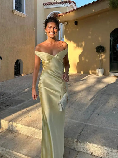 Jeune femme souriante en robe de soirée jaune élégante, tenant un sac à main, posant devant une maison méditerranéenne au coucher du soleil.