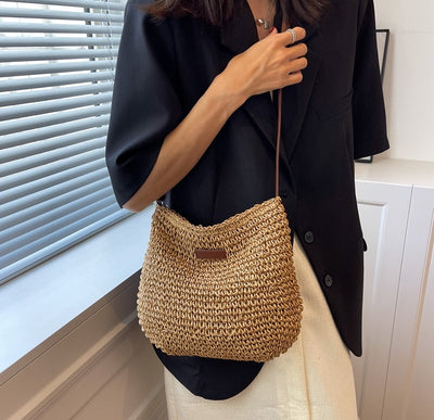 Femme portant un sac en osier tressé, tenue élégante avec veste noire et jupe blanche, près d'une fenêtre. Mode chic, accessoires tendance.