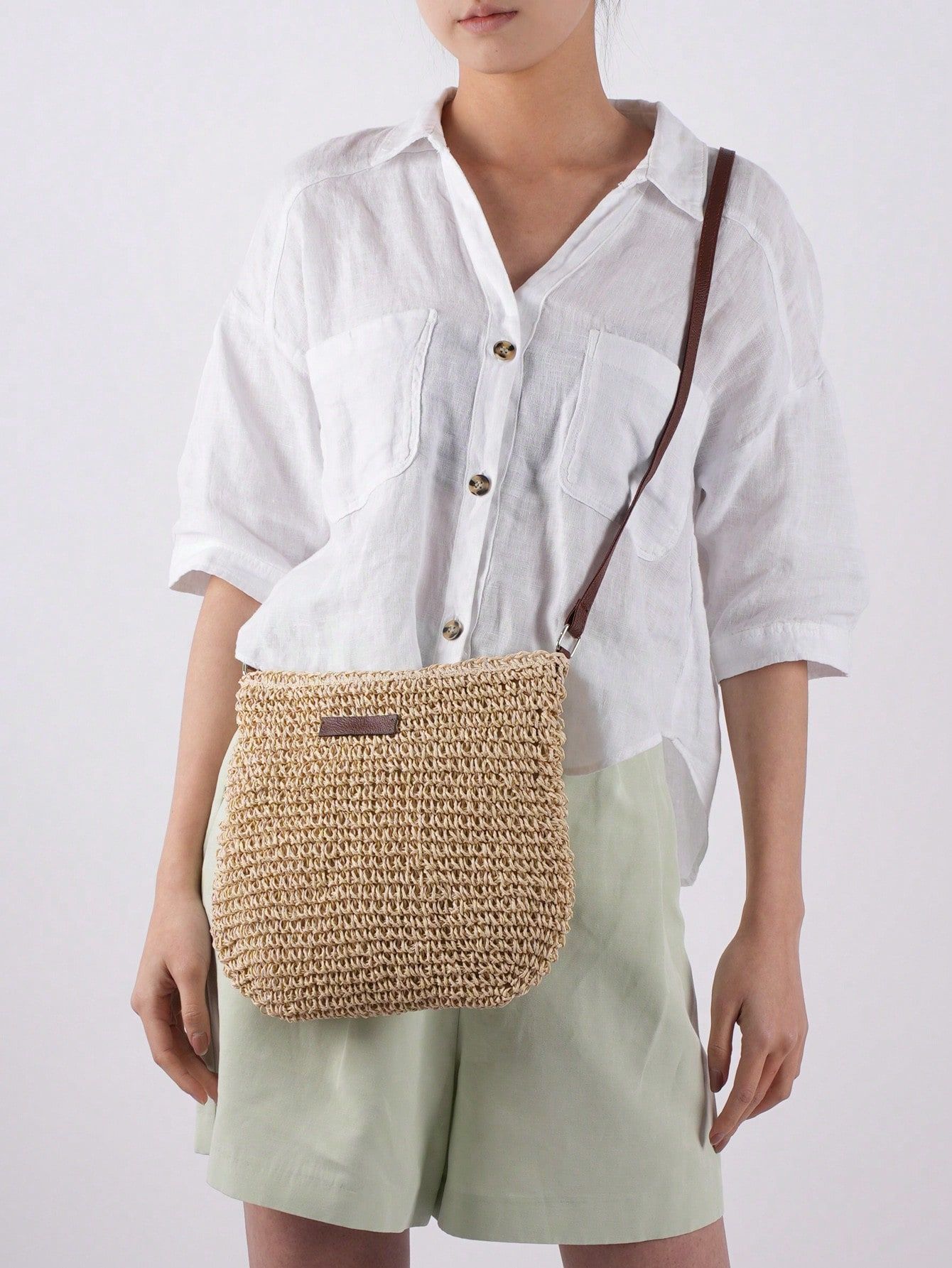 Femme portant un sac en paille tressée avec bandoulière marron, chemise blanche en lin et short vert clair. Mode estivale, accessoires tendance.
