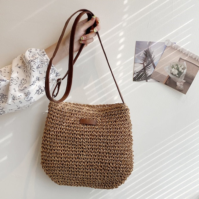 Sac à main en paille tressée avec bandoulière marron, tenu par une main. Décor mural minimaliste avec photos. Mode éthique, accessoire d'été.