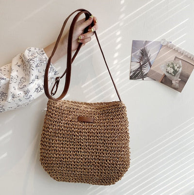 Sac à main en paille tressée avec bandoulière marron, tenu par une main. Décor mural minimaliste avec photos. Mode éthique, accessoire d'été.
