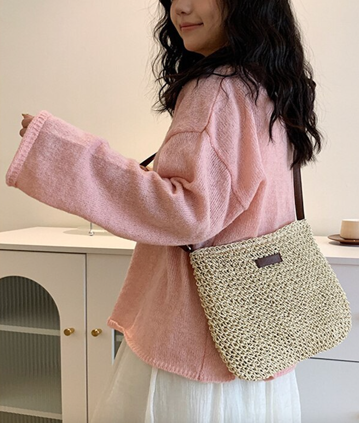 Femme portant un pull rose et un sac en paille tressée, debout devant un meuble blanc. Mode décontractée, accessoires tendance.