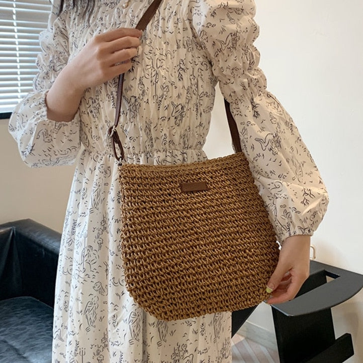 Femme en robe blanche à motifs, portant un sac en paille tressée marron avec bandoulière en cuir, dans un intérieur moderne.