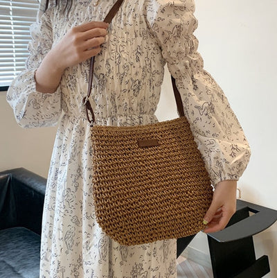Femme en robe blanche à motifs, portant un sac en paille tressée marron avec bandoulière en cuir, dans un intérieur moderne.