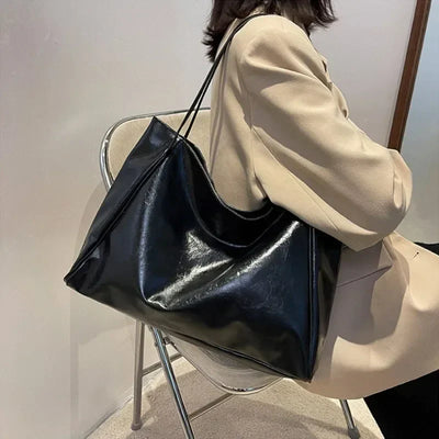 Sac à main noir en cuir brillant porté par une femme en manteau beige, assise sur une chaise moderne. Accessoire mode élégant et tendance.