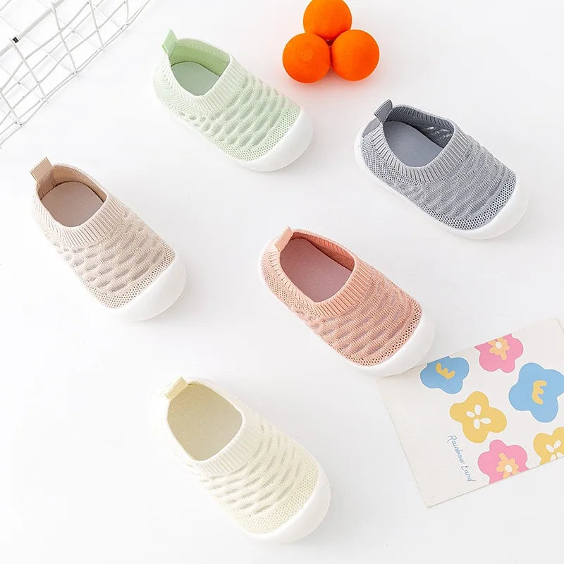 Cinq chaussons bébé en tricot de différentes couleurs sur fond blanc, avec des motifs floraux et des oranges en arrière-plan.