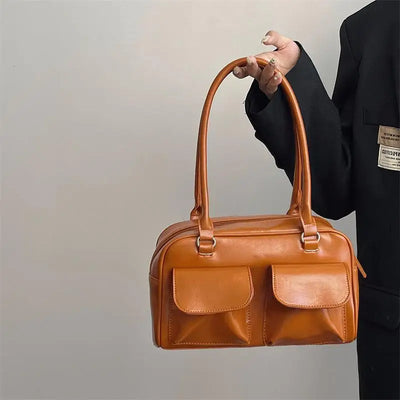 Sac à main en cuir marron avec deux poches avant, porté par une personne en veste noire. Accessoire mode élégant et tendance.