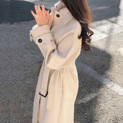 Femme portant un manteau beige élégant, ceinturé, manches longues, sur fond de rue ensoleillée. Mode automne, style urbain chic.