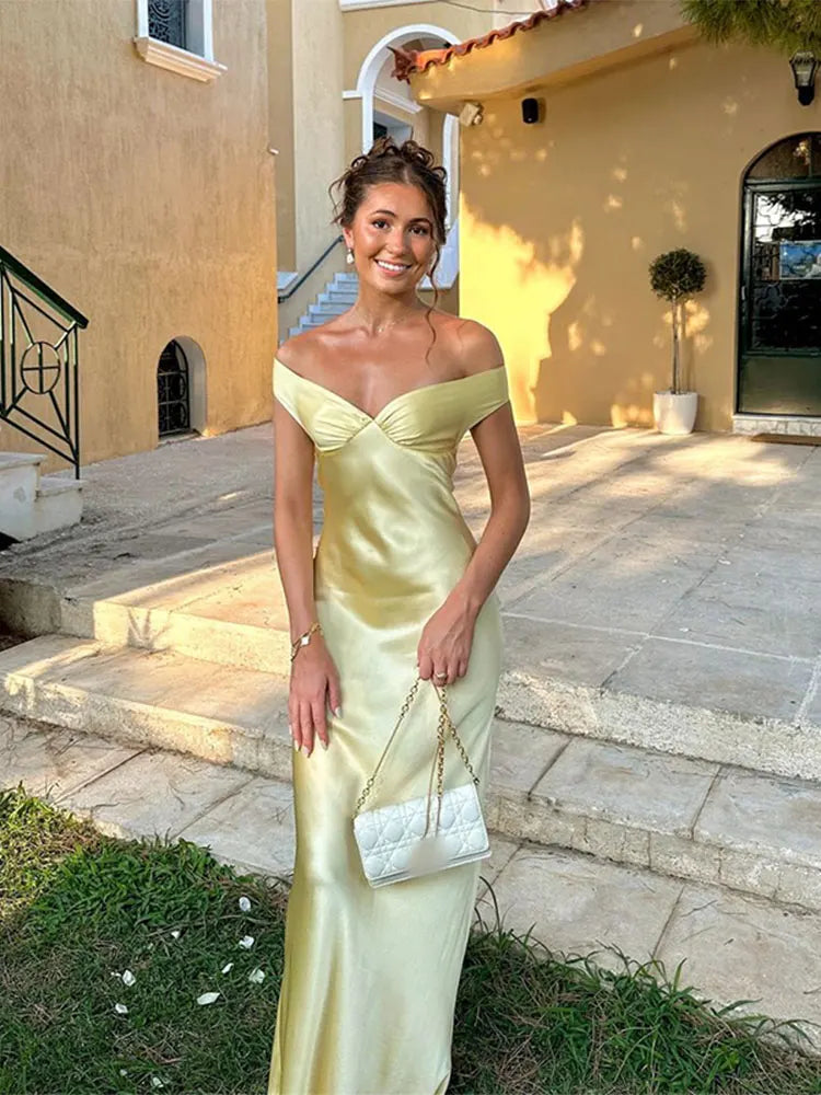 Jeune femme en robe de soirée jaune élégante, posant devant une maison méditerranéenne. Elle tient un sac à main blanc. Style chic et estival.