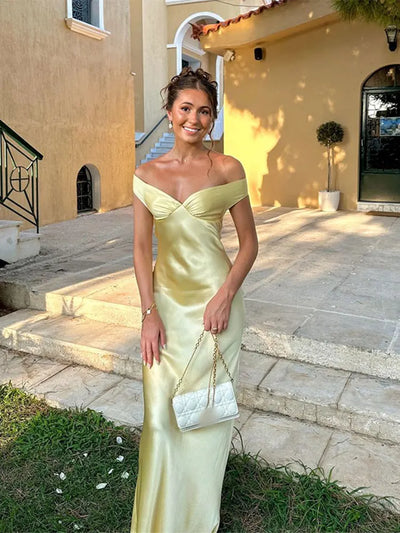 Jeune femme en robe de soirée jaune élégante, posant devant une maison méditerranéenne. Elle tient un sac à main blanc. Style chic et estival.