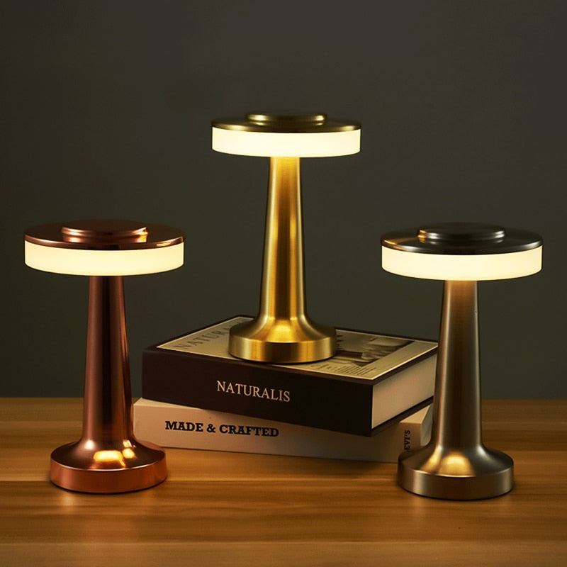 Trois lampes de table LED modernes en métal doré, cuivre et argent, posées sur des livres, éclairant un bureau en bois. Éclairage intérieur design.