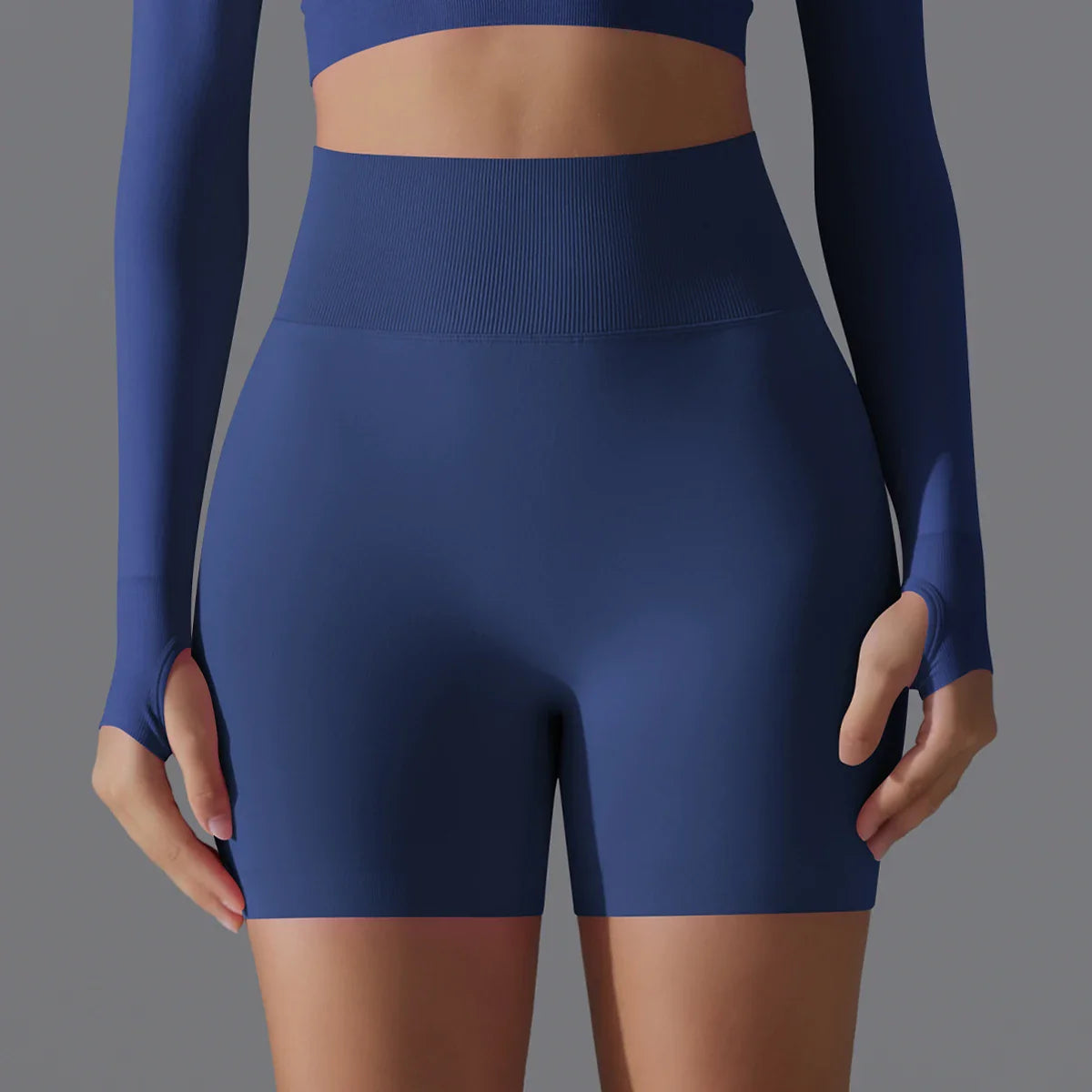 Femme en tenue de sport bleue, portant un short taille haute et un haut à manches longues, sur fond gris. Mode fitness, vêtements de sport élégants.