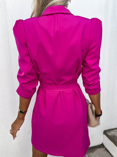 Femme en robe blazer rose vif, manches longues, ceinture à la taille, montre au poignet, sac beige à la main, fond blanc.