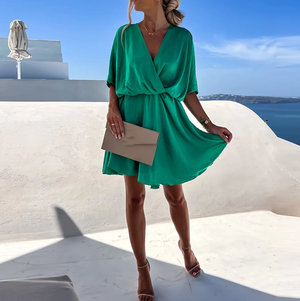 Femme en robe verte élégante, tenant une pochette beige, posant sur une terrasse ensoleillée avec vue sur la mer Égée, ciel bleu clair.