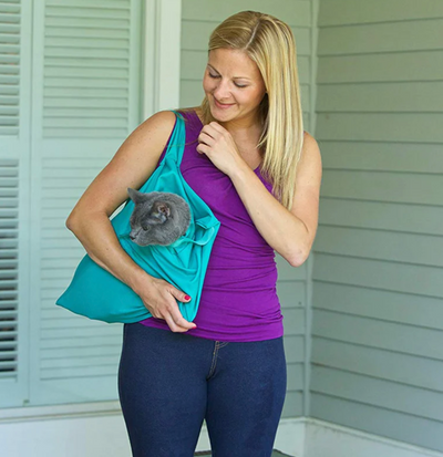 Femme souriante portant un chat gris dans un sac vert, debout devant une maison. Accessoire pratique pour animaux de compagnie.