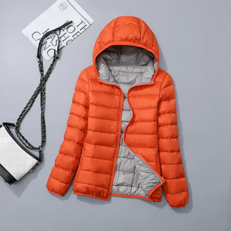 Doudoune orange à capuche, doublure grise, posée à plat avec sac à main blanc et noir en chaîne, fond gris. Mode hiver, vêtements chauds.