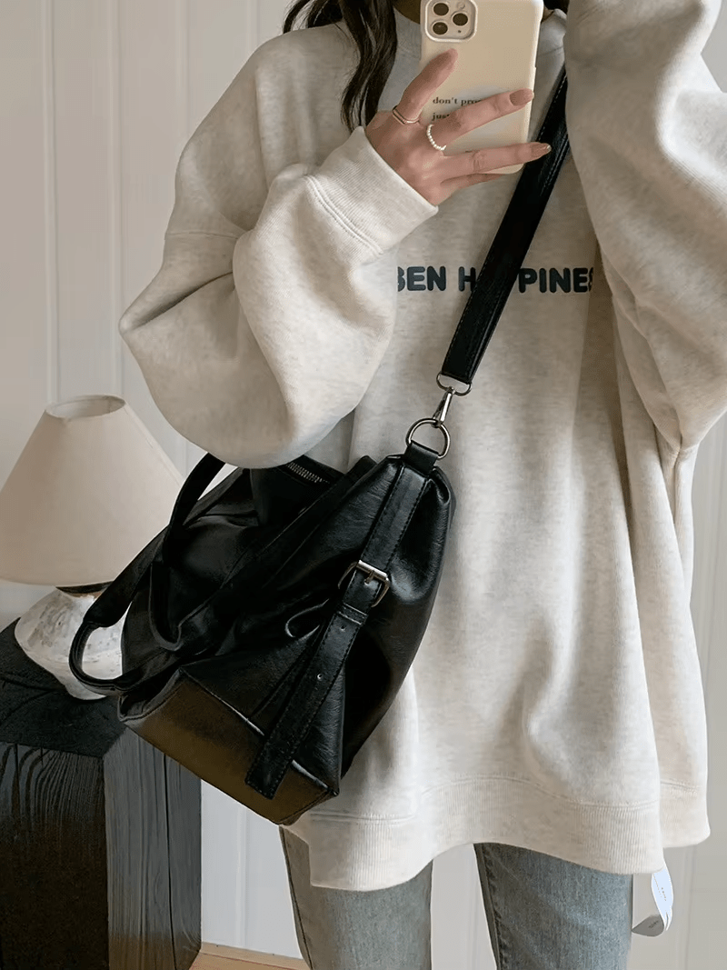 Femme portant un sweat-shirt gris et un sac à main noir en cuir, tenant un smartphone. Mode décontractée, accessoires tendance, style urbain.