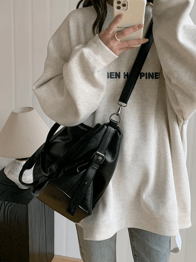 Femme portant un sweat-shirt gris et un sac à main noir en cuir, tenant un smartphone. Mode décontractée, accessoires tendance, style urbain.