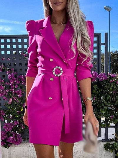 Robe blazer rose vif pour femme avec ceinture ornée, manches longues et boutons dorés, devant un fond fleuri. Mode élégante et tendance.