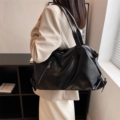 Femme en manteau beige portant un grand sac à main noir en cuir, debout près d'une étagère noire. Mode élégante, accessoires tendance.