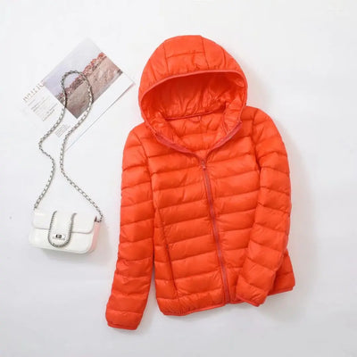 Doudoune orange à capuche posée à plat, accompagnée d'un sac à main blanc avec chaîne argentée et magazine. Mode automne-hiver tendance.