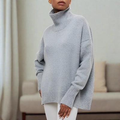Femme portant un pull gris à col roulé, manches longues, dans un salon moderne. Mode automne-hiver, confort et élégance.