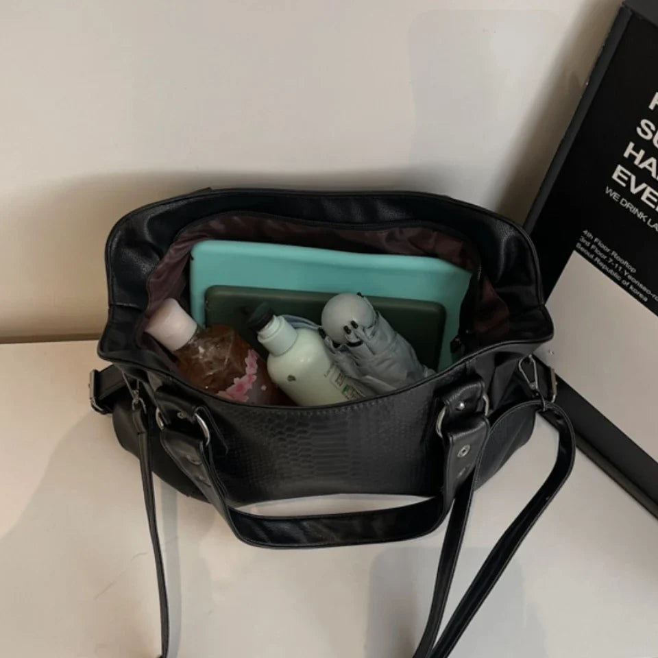 Sac à main noir ouvert avec tablette, bouteille d'eau, crème et peluche à l'intérieur, posé sur une table blanche. Accessoires de mode et technologie.