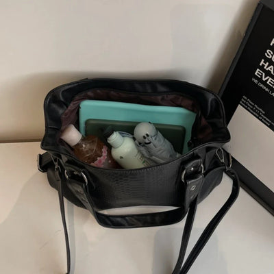 Sac à main noir ouvert avec tablette, bouteille d'eau, crème et peluche à l'intérieur, posé sur une table blanche. Accessoires de mode et technologie.