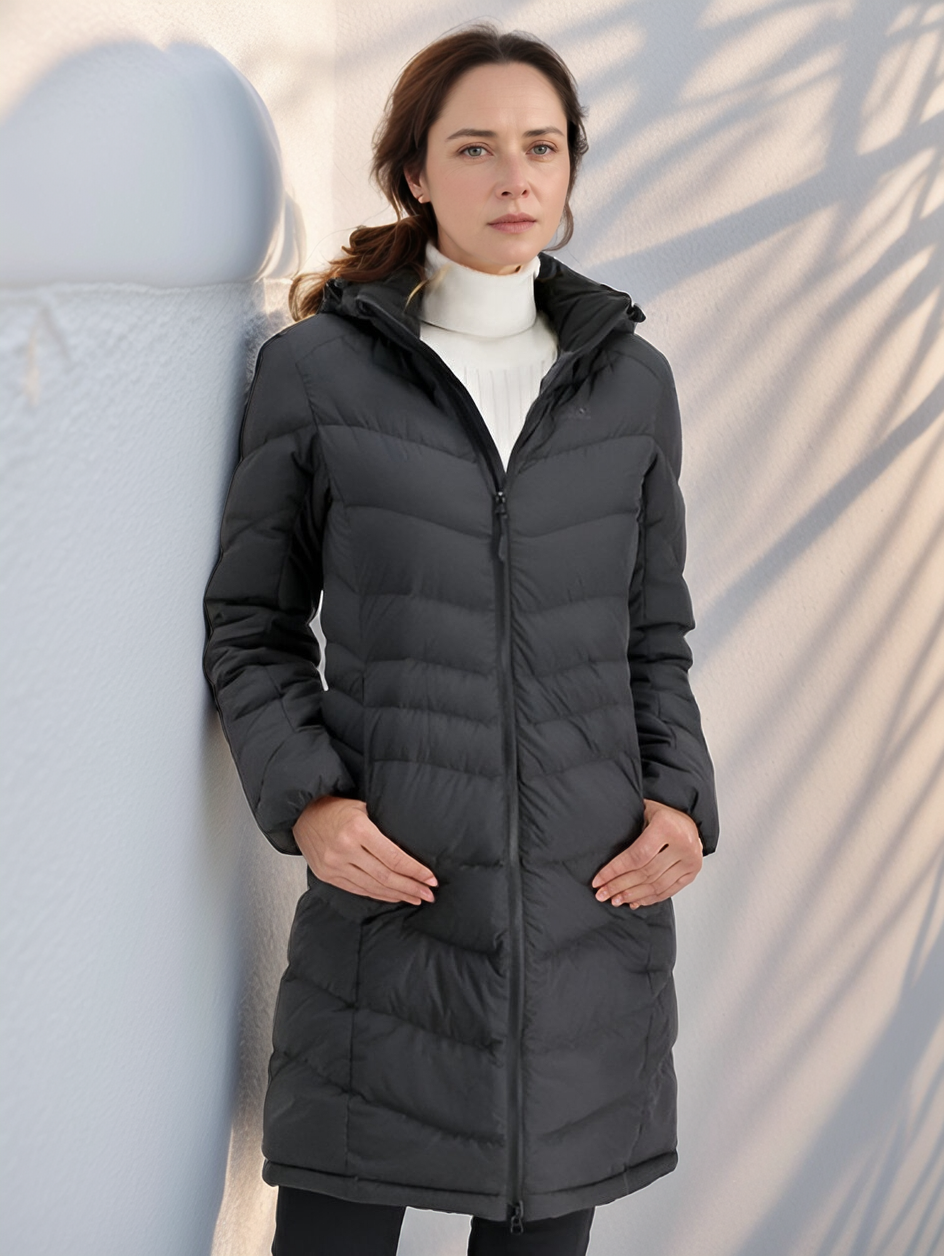 Femme portant un manteau d'hiver noir matelassé, appuyée contre un mur blanc, lumière douce et ombres de branches. Mode hiver élégante et moderne.