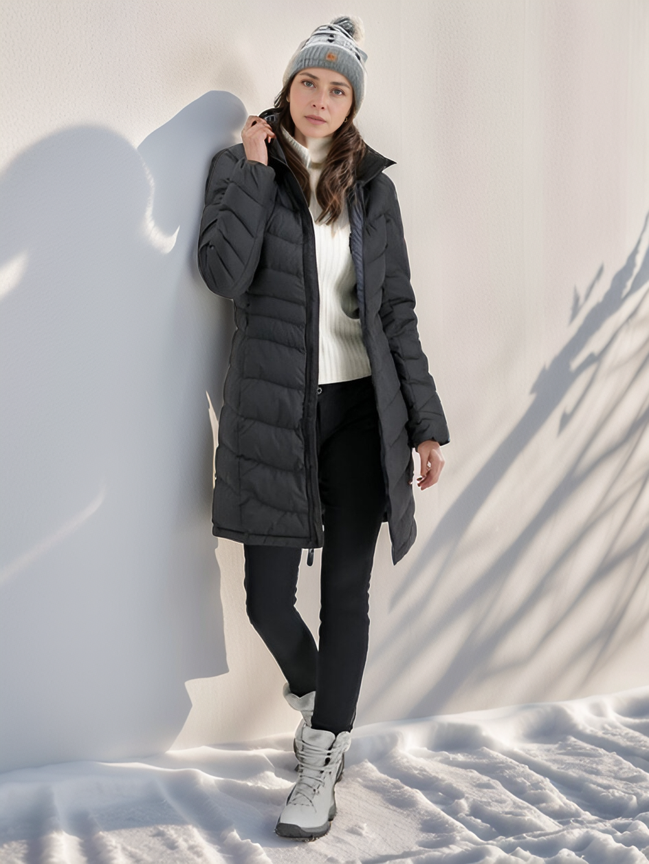 Femme en manteau d'hiver noir, bonnet gris, pose contre un mur enneigé. Mode hiver, vêtements chauds, style décontracté, extérieur neigeux.