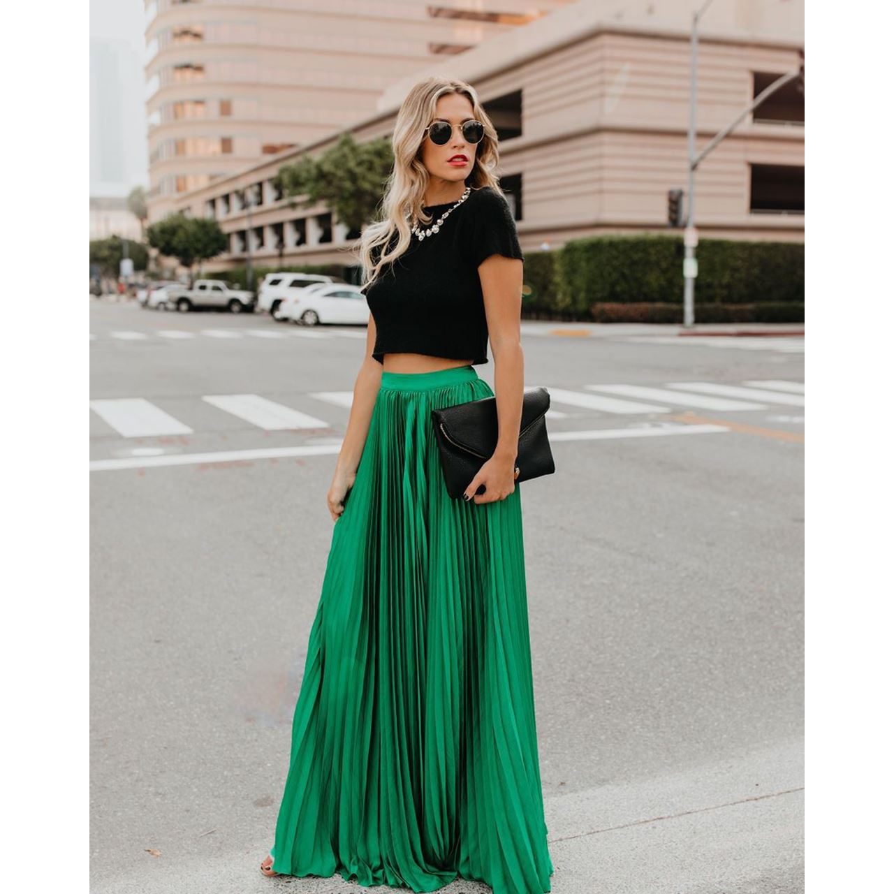 Femme élégante en jupe longue verte et haut noir, tenant une pochette noire, traverse une rue urbaine. Mode chic, style urbain, tenue tendance.