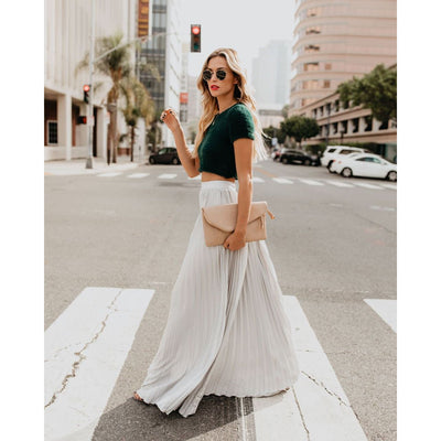 Femme élégante traversant une rue urbaine, portant un haut vert et une jupe longue blanche, tenant une pochette beige. Mode urbaine chic.