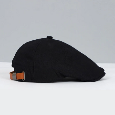 Casquette plate noire en tissu avec sangle réglable en cuir marron, posée sur une surface blanche, fond gris. Accessoire mode élégant et classique.