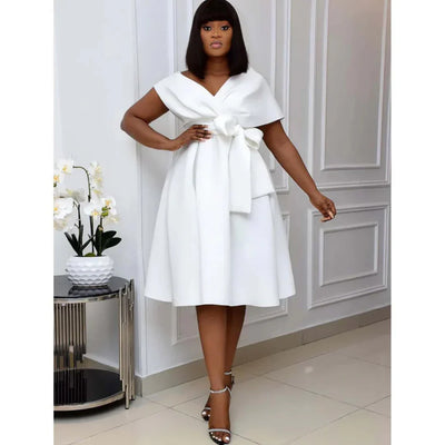 Femme élégante en robe blanche avec nœud, posant près d'une table moderne et orchidées blanches, dans un intérieur chic et minimaliste.