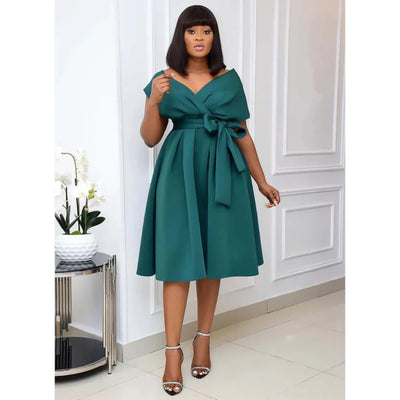 Robe verte élégante avec ceinture nouée, femme debout dans un intérieur moderne, orchidées blanches en arrière-plan, style chic et sophistiqué.