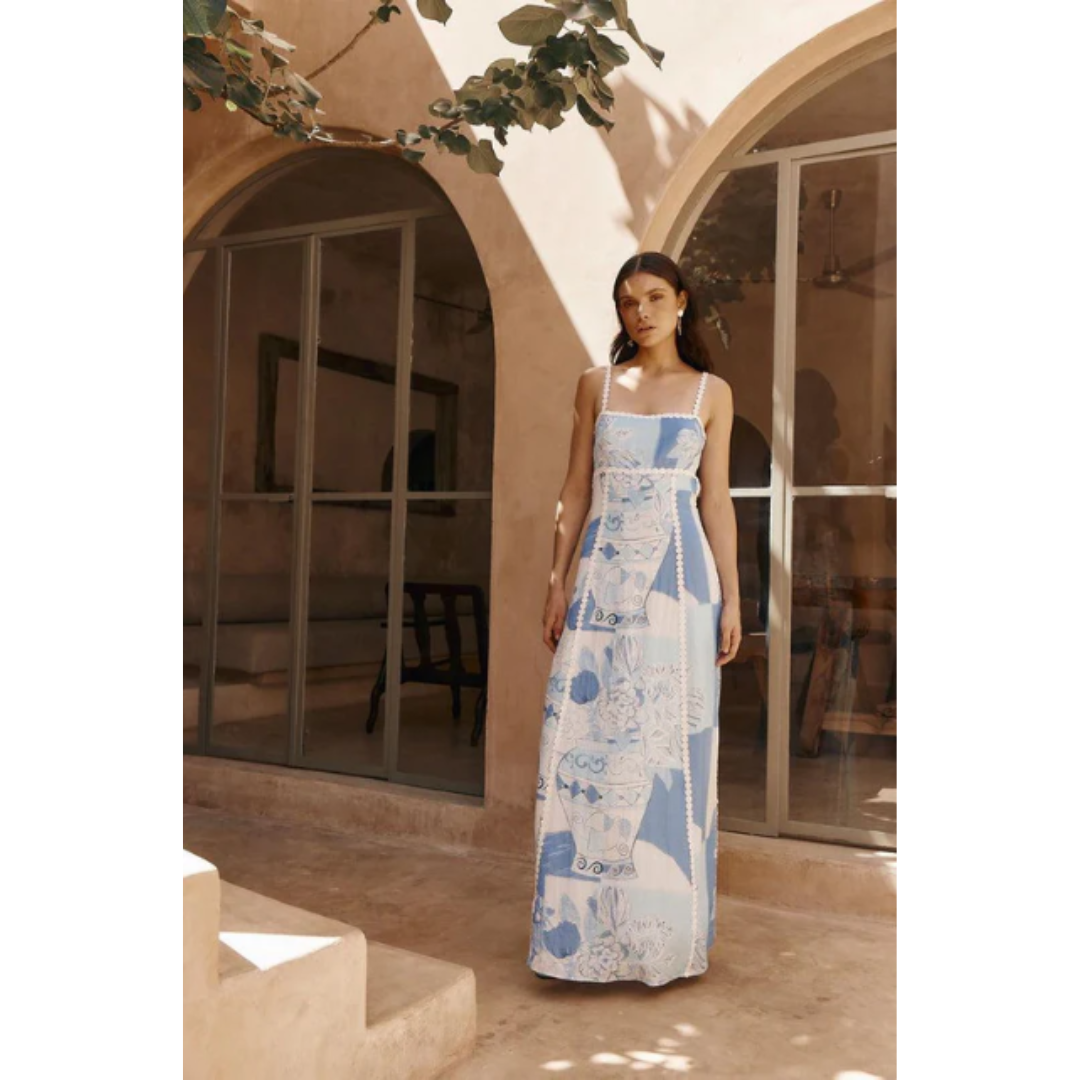 Robe longue d'été bleue et blanche à motifs géométriques, portée par une femme devant une maison méditerranéenne avec arches et plantes.