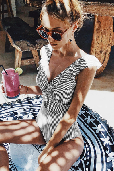 Femme en maillot de bain rayé, lunettes de soleil, buvant un smoothie rose avec une rondelle de citron vert, assise sur un tapis rond à motifs.